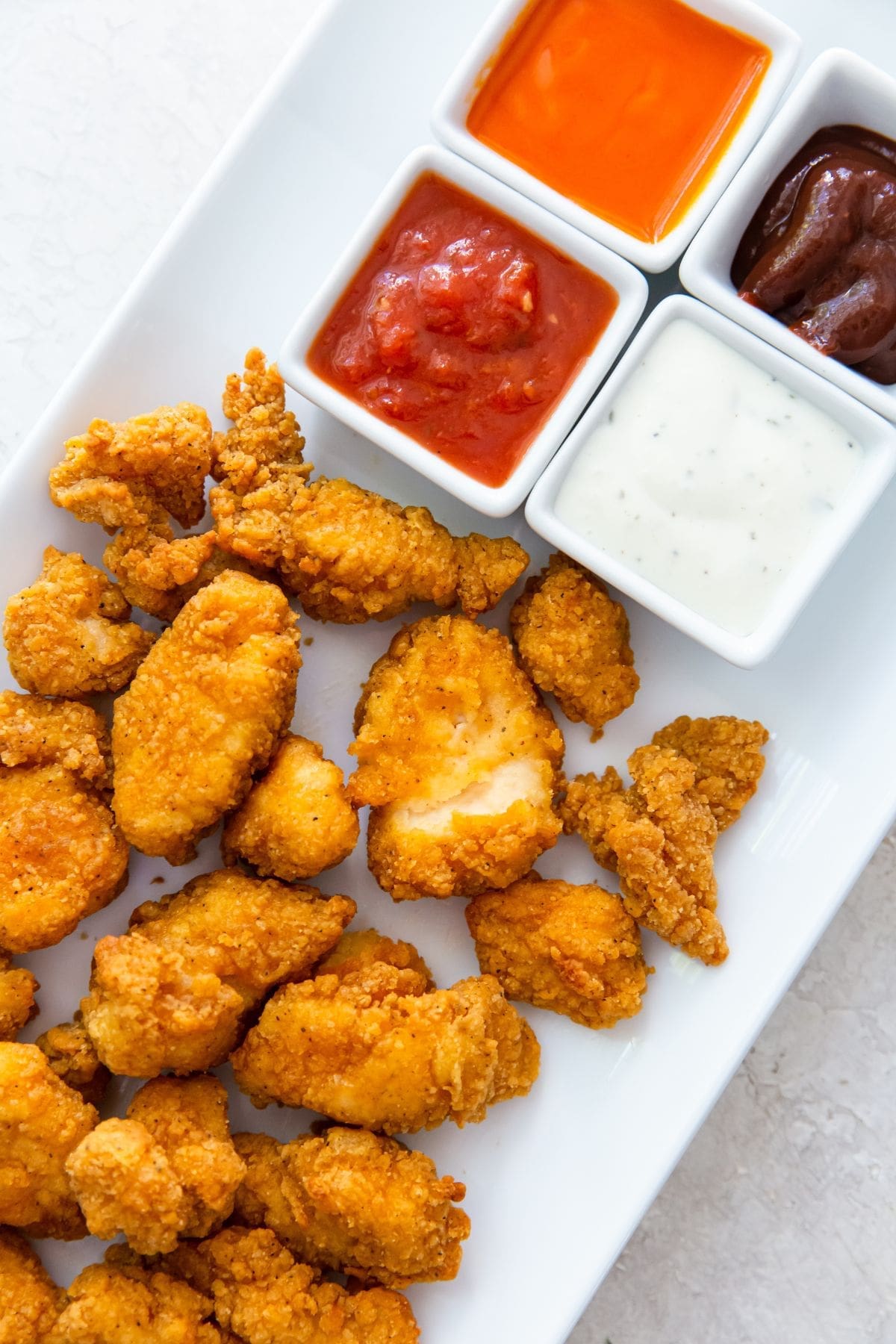 white plate with popcorn chicken on it buffalo, ranch, bbq, and ketchup in the back