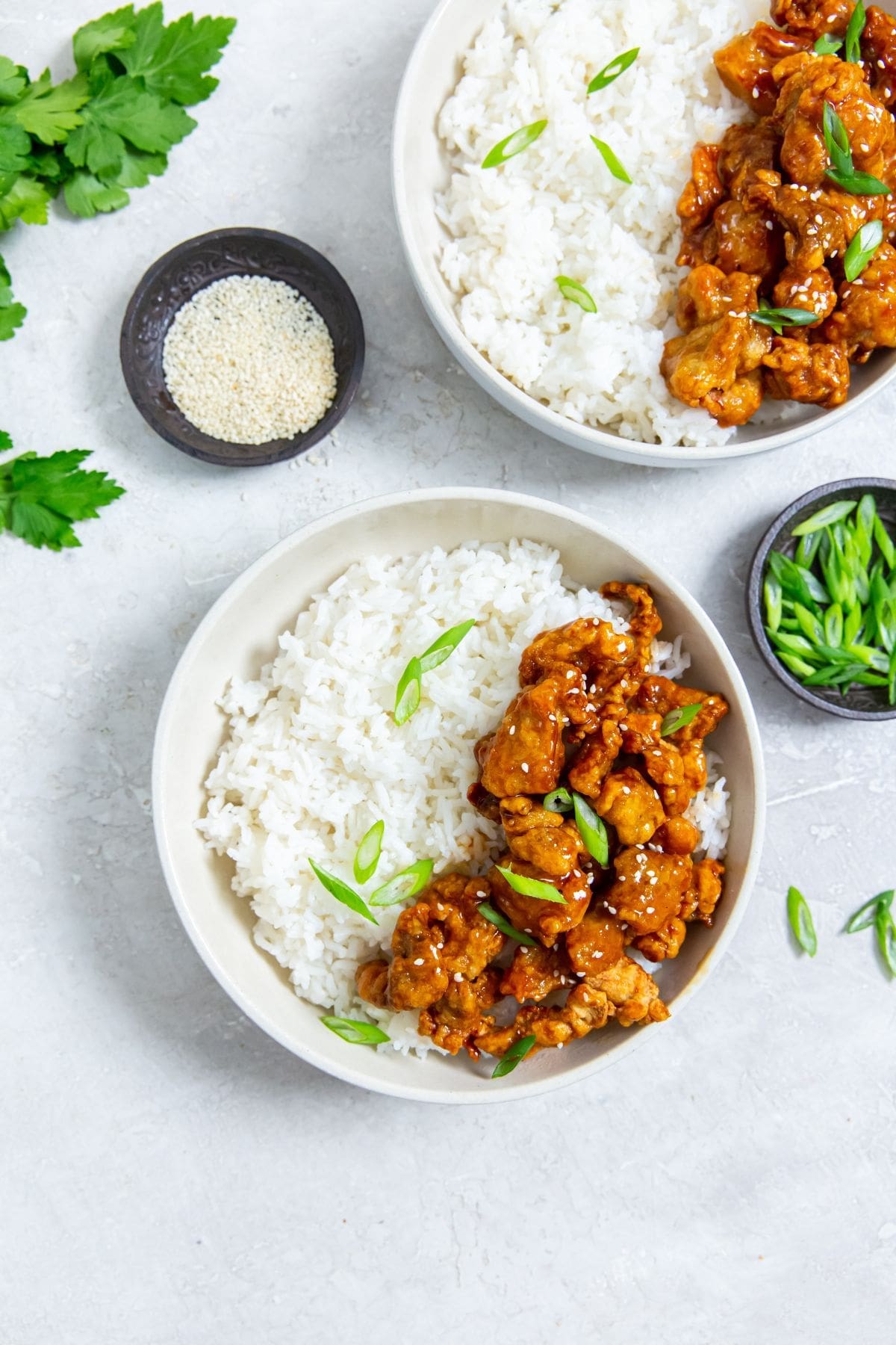 white bowl with chicken and rice inside. scallions on top. Gray towel next to it. scallions and sesame seeds in the back. chopsticks on the bowl