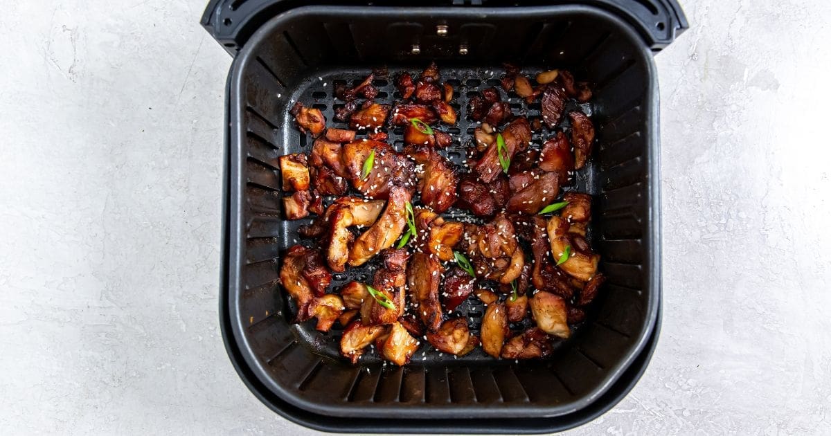 Black air fryer with cooked chicken tossed in sauce and sprinkled with sesame seeds and scallions