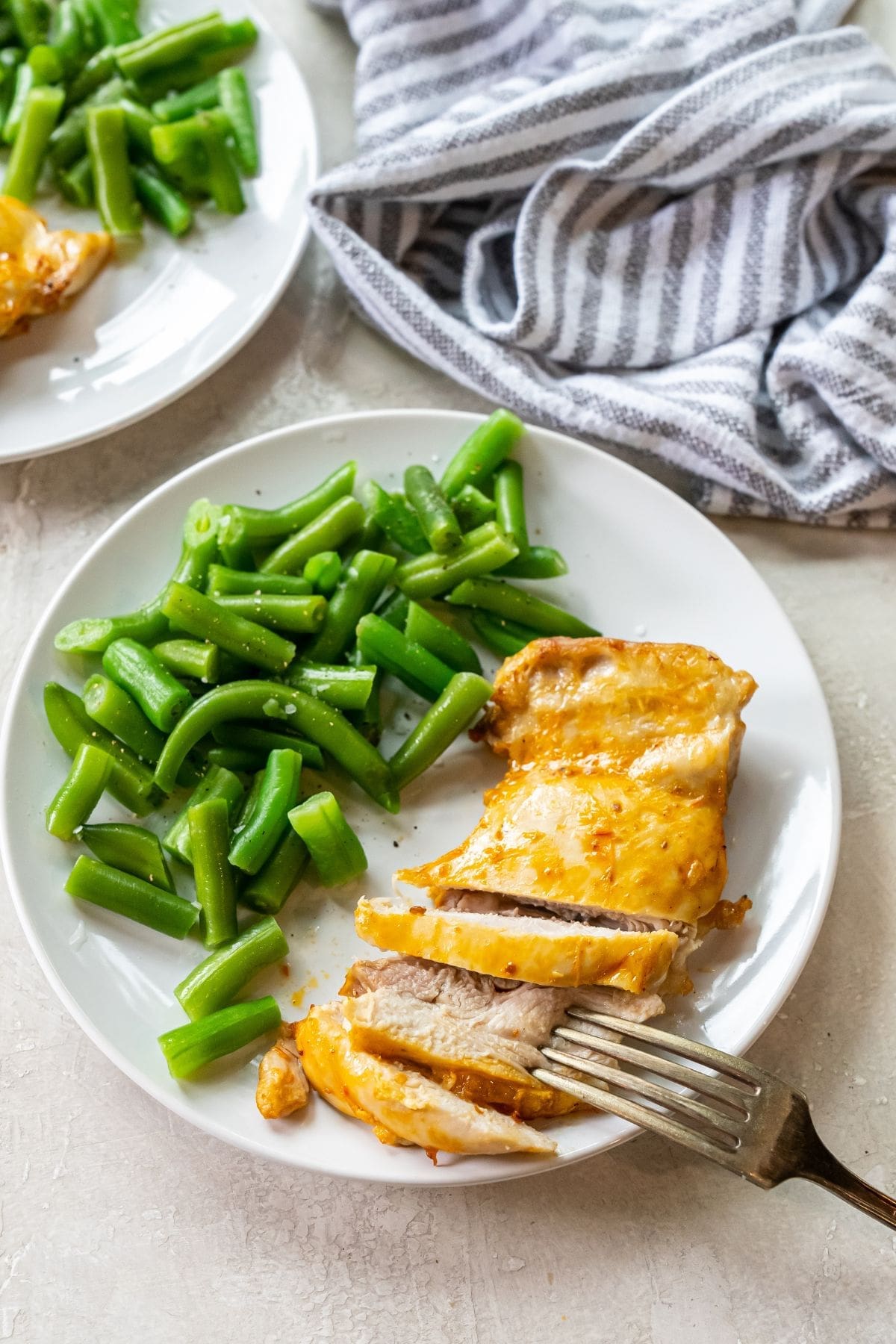 white plate with chicken and green beans on it. fork next to chicken. striped towel next to plates.