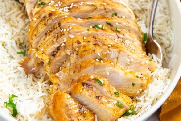 Sliced roasted chicken breast served over a bed of white rice garnished with sesame seeds and chopped herbs in a white bowl.